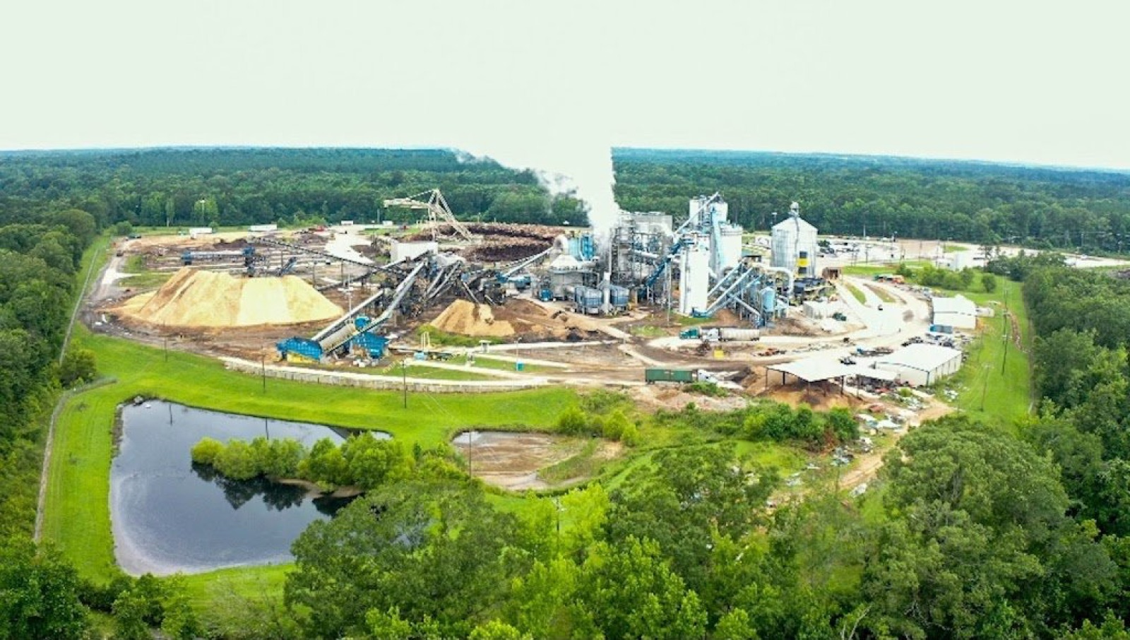 Aerial view of the Drax wood pellet plant in Gloster, Mississippi, with log piles and a steam plume, surrounded by forest